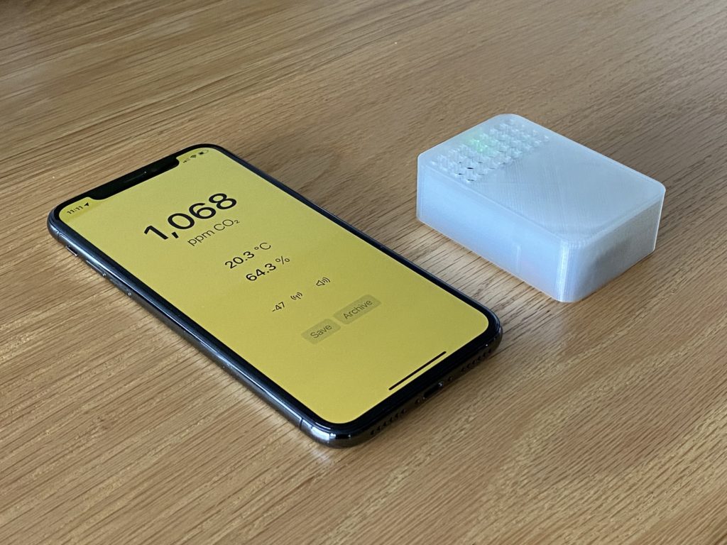 A photo of the CO2 sensor in a 3D printed case, next to an iPhone X running the iOS app to record readings and locations.